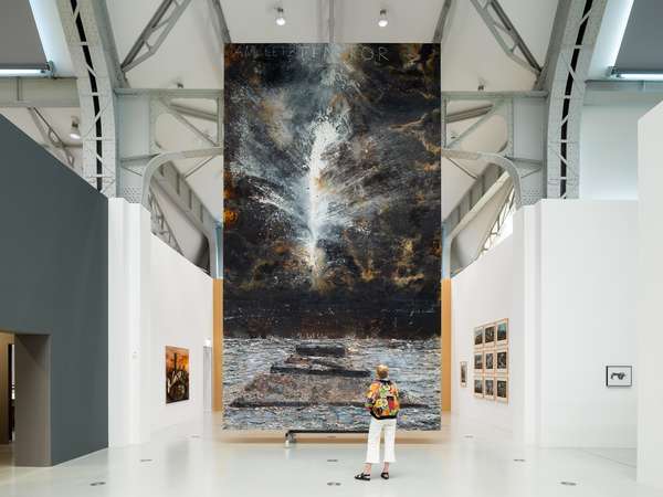 Eine Frau steht in einer großen Ausstellungshalle vor einem ebenso riesigen Gemälde, das im Hochformat eine abstrakte Landschaft zeigt.