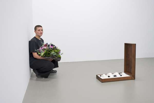 Eine junge Frau mit geschorenen Haaren hockt an eine Wand gelehnt mit einem Blumenstrauß in der Hand. Neben ihr steht ein geöffneter Holzkoffer mit weißen Glasobjekten.