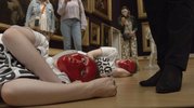 In einem Museum mit Bildern an den Wänden liegen zwei Personen mit jeweils einer roten Gesichtsmaske aus Plastik auf dem Boden. Der Bildausschnitt ist sehr nah gewählt. Weitere Personen stehen um die Liegenden drum herum.