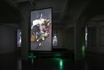 Eine Fotografie von dem Ausstellungsraum - in der Mitte des Raumes hängt ein länglicher Bildschirm worauf projiziert Menschen auf dem Boden zu erkennen sind, ineinander verschlungen im schicken Kostüm.  