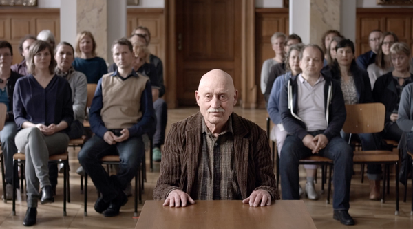 The film still from "Forcing Satan to Do This" shows an elderly man in a close-up. Behind him, people are sitting in a courtroom. They can only be seen in a blur.