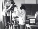 Ein Mann an einer Kamera steht an der linken Bildhälfte. Eine Frau vor ihm korrigiert die Zoomeinstellung der Linse per Hand an der Kamera und schaut gleichzeitig auf einen Bildschirm, der rechts neben ihr auf einem Tisch steht. Sie befinden sich in einem Klassenzimmer.