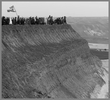 Eine schwarz/weiß Fotografie von einem steilen Hang worauf eine Gruppe von Menschen stehen mit einer Flagge. Im Hintergrund sind weitere Hügel zu sehen.