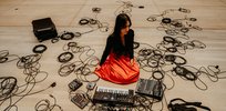 Eine Fotografie der Musikerin: Sie sitzt am Boden mit orangem Kleid, viele Kabel liegen um sie herum, zwei Synthesizer liegen vor ihr