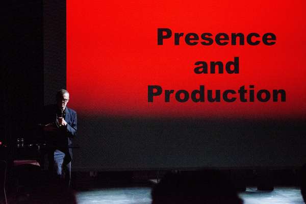 Auf einer großen roten Leinwandprojektion steht in schwarzen Lettern "Presence and Production". Ein Mann mittleren Alters steht an der linken Bildhälfte, leicht angestrahlt mit einem Mikro in der Hand. Er schaut in die Kamera.