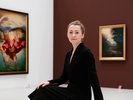 Eine Frau im schwarzen langen Kleid sitzt in gedrehter Position in einer Gemäldegallerie und schaut den Betrachter direkt an. Sie trägt ihre asch-blonden Haare zu einem Dutt und lächelt leicht.