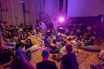 Viele Studierende sitzen in der Aula auf dem Boden. Zwei Künstler*innen performen in der Mitte des Raumes. Der Raum ist in lila Licht getaucht.