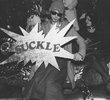 Ein verkleideter Mann mit Sonnenbrille hält ein Schild in Sternform in die Kamera. Darauf steht "Suckle". Das Bild ist in Schwarz-Weiß aufgenommen.
