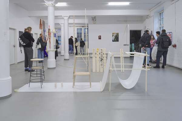 Menschen stehen in einem Ausstellungsraum und betrachten Werke und Installationen. Eine Installation dominiert die Mitte des Raumes. Sie besteht aus mehreren schlanken Holz-Elementen.