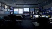 In einem dunklen Raum ohne Fenster sind eine Vielzahl an eingeschalteten Monitoren zu sehen, die auf Tischen stehen oder an Wänden hängen.