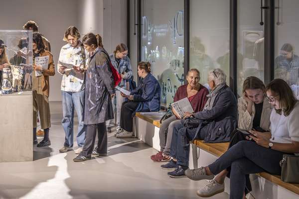 Viele Besucher*innen halten sich im Ausstellungsraum auf. Manche sitzen im großen Fenster des Raumes, andere stehen. Viele schauen sich die Exponate und auch die Texte an den Wänden an.