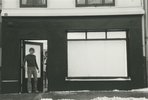 Eine Häuserfront ist zu sehen. Auf der rechten Seite ein großes Schaufenster, das weiß verhangen wurde. Auf der linken Seite öffnet sich eine Ladentür. Ein Mann steht im Türrahmen.