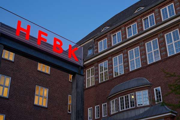 Auf der linken Bildhälfte ist die Wortbildmarke in leuchtenden roten Lettern der HFBK zu lesen. Im Hintergrund erstreckt sich die Gebäudefassade der HFBK.