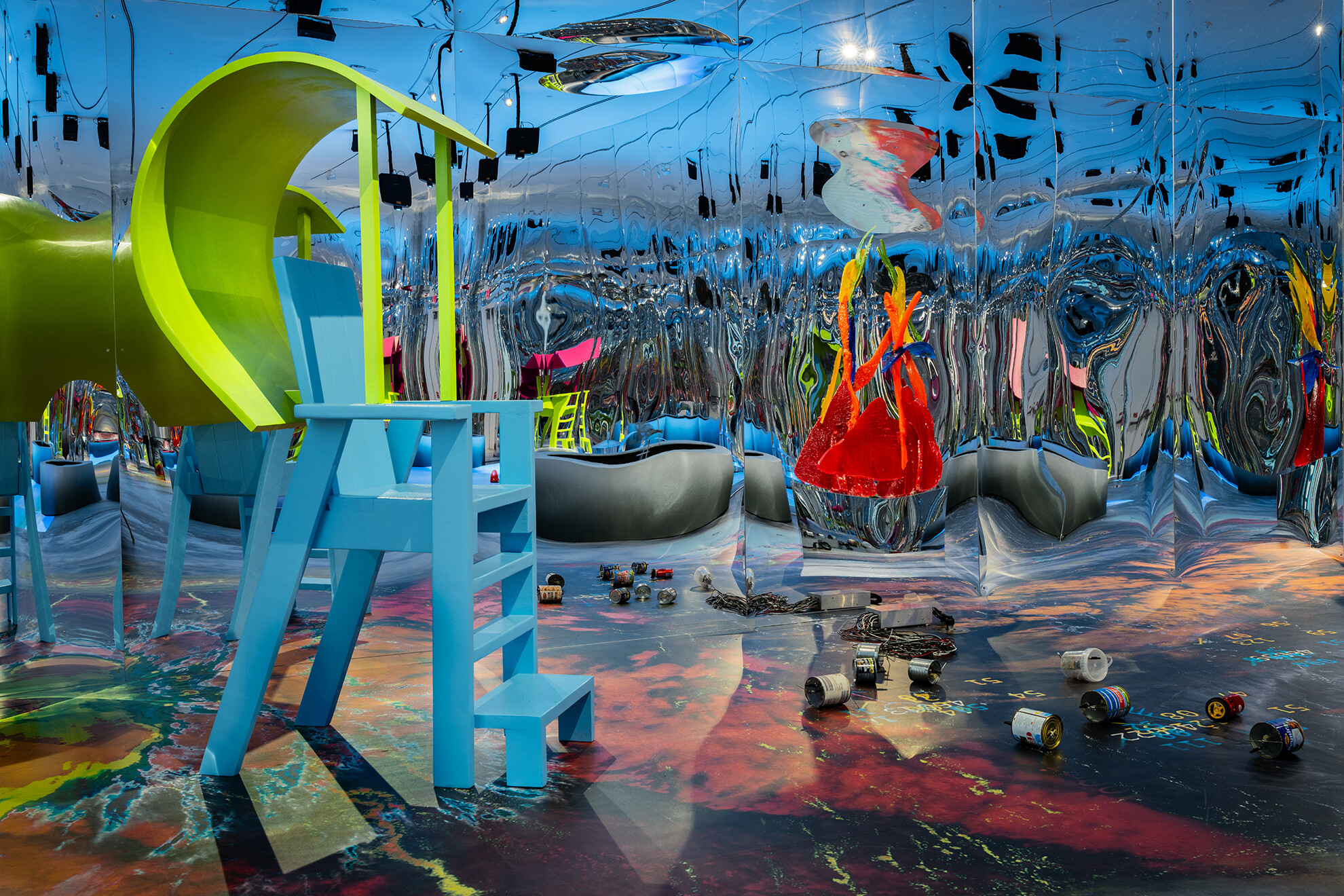 Colorful, immersive installation artwork in a room with highly reflective mirrored walls. A large light-blue high-chair-like structure stands in the foreground next to a curved neon-green form. Bright red, organic shapes hang in the space, and paint cans and cables are scattered across the vividly painted floor. The mirrors create multiple reflections and spatial distortions.