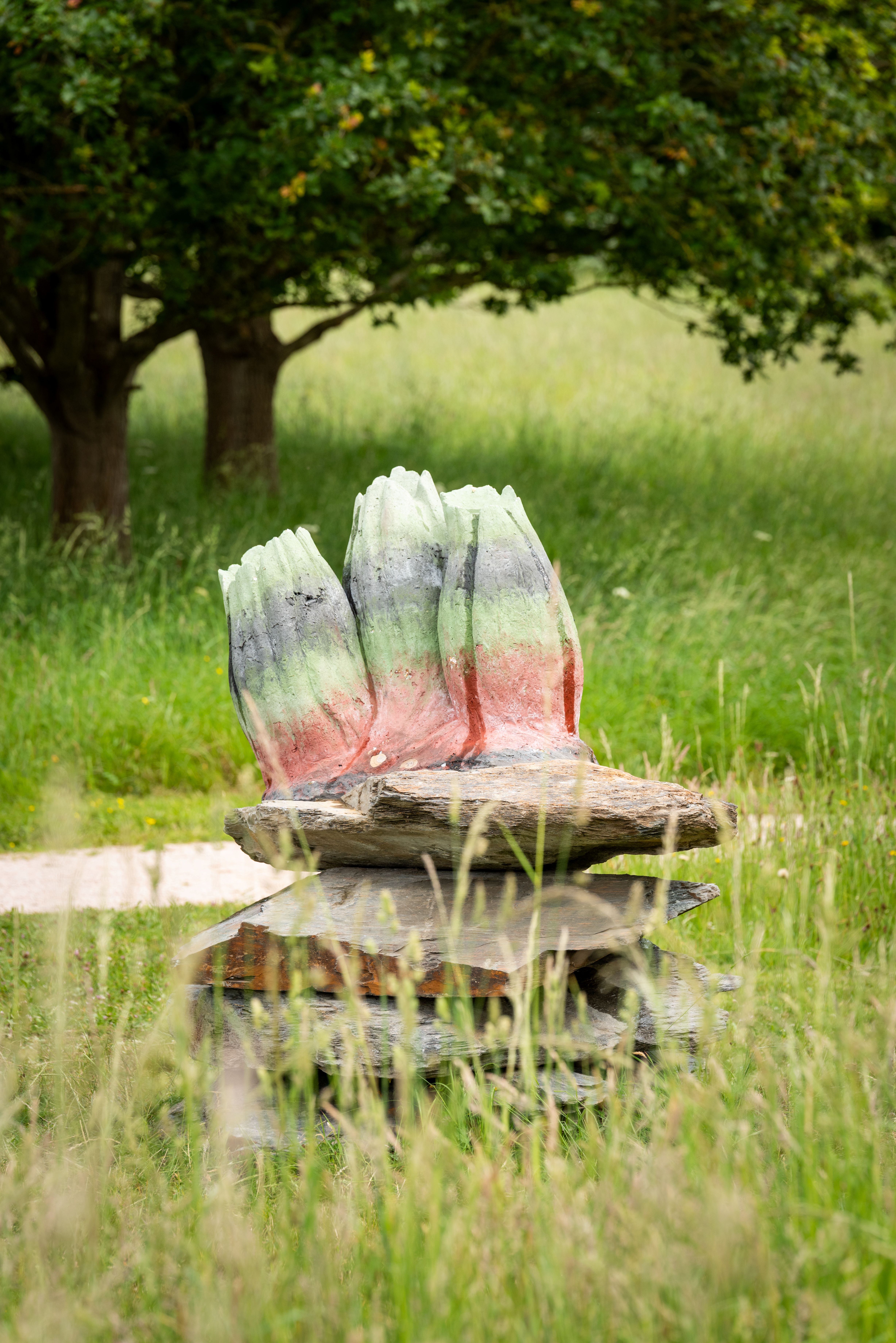 Organically shaped sculpture with a green, gray, and reddish surface placed on stacked wooden slabs. It stands in a grassy meadow with tall grass, and a tree is visible in the background.