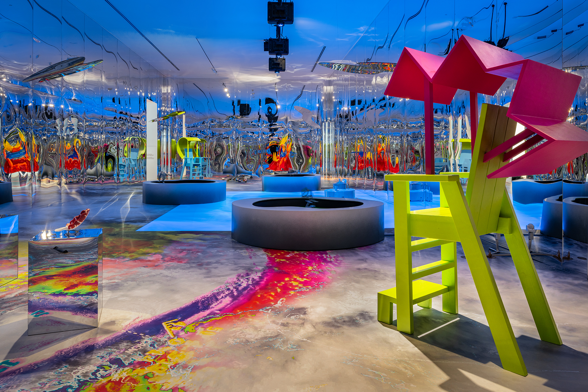 Large-scale, vividly colored installation in a room with fully mirrored walls. Several sculptural furniture-like objects in neon colors—including a pink and green high-chair-like structure—stand on a floor painted with flowing, multicolored patterns. Circular platforms and a round basin structure the space, while the mirrors multiply and distort the forms through reflections.