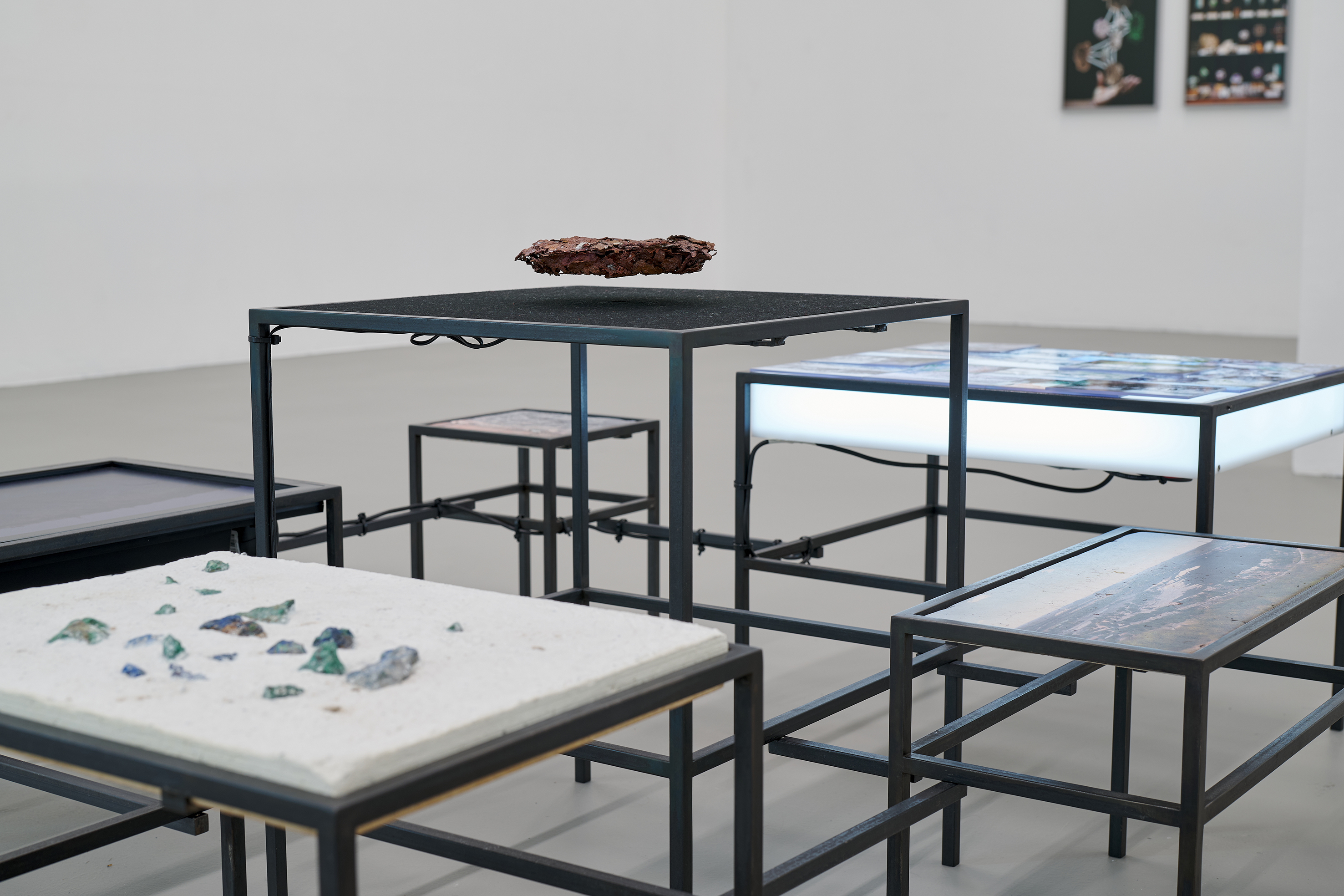An installation composed of black metal tables of varying heights is arranged within a white gallery space. The tabletops display different materials and objects, including glass fragments, photographic surfaces, and a dark form appearing to hover above a central table. Some surfaces are illuminated from below, creating a scientific and archival atmosphere. The arrangement resembles a laboratory or research station in which diverse materials are brought into relation with one another.