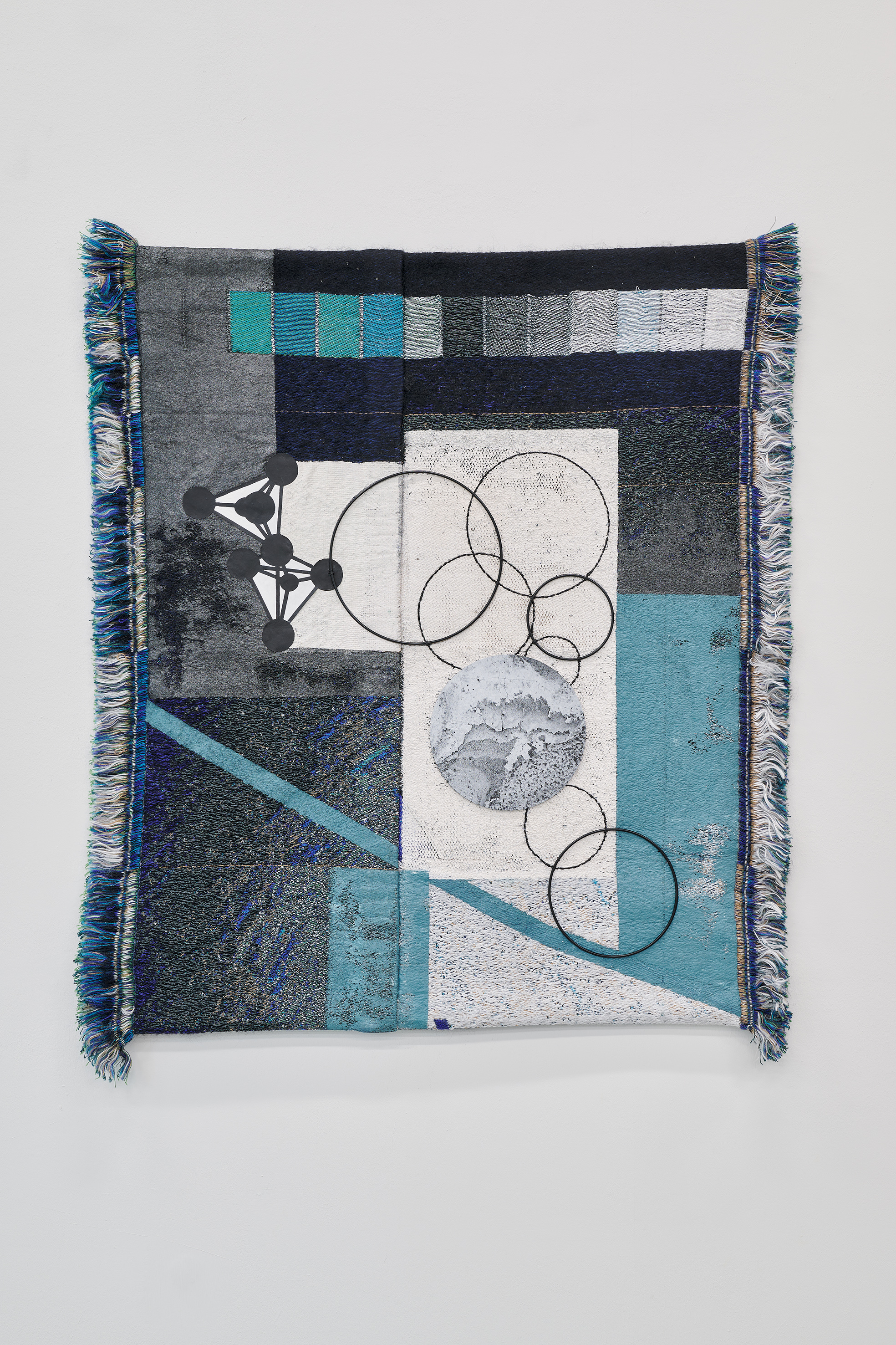 A square textile artwork hangs on a white wall. The woven composition combines abstract geometric forms in shades of blue, grey, black, and white with graphic elements such as circles, lines, and network-like structures. Different weaving techniques and materials create a richly layered surface. Fringed edges emphasize the handcrafted quality of the piece, contrasting with its precise and constructive composition.