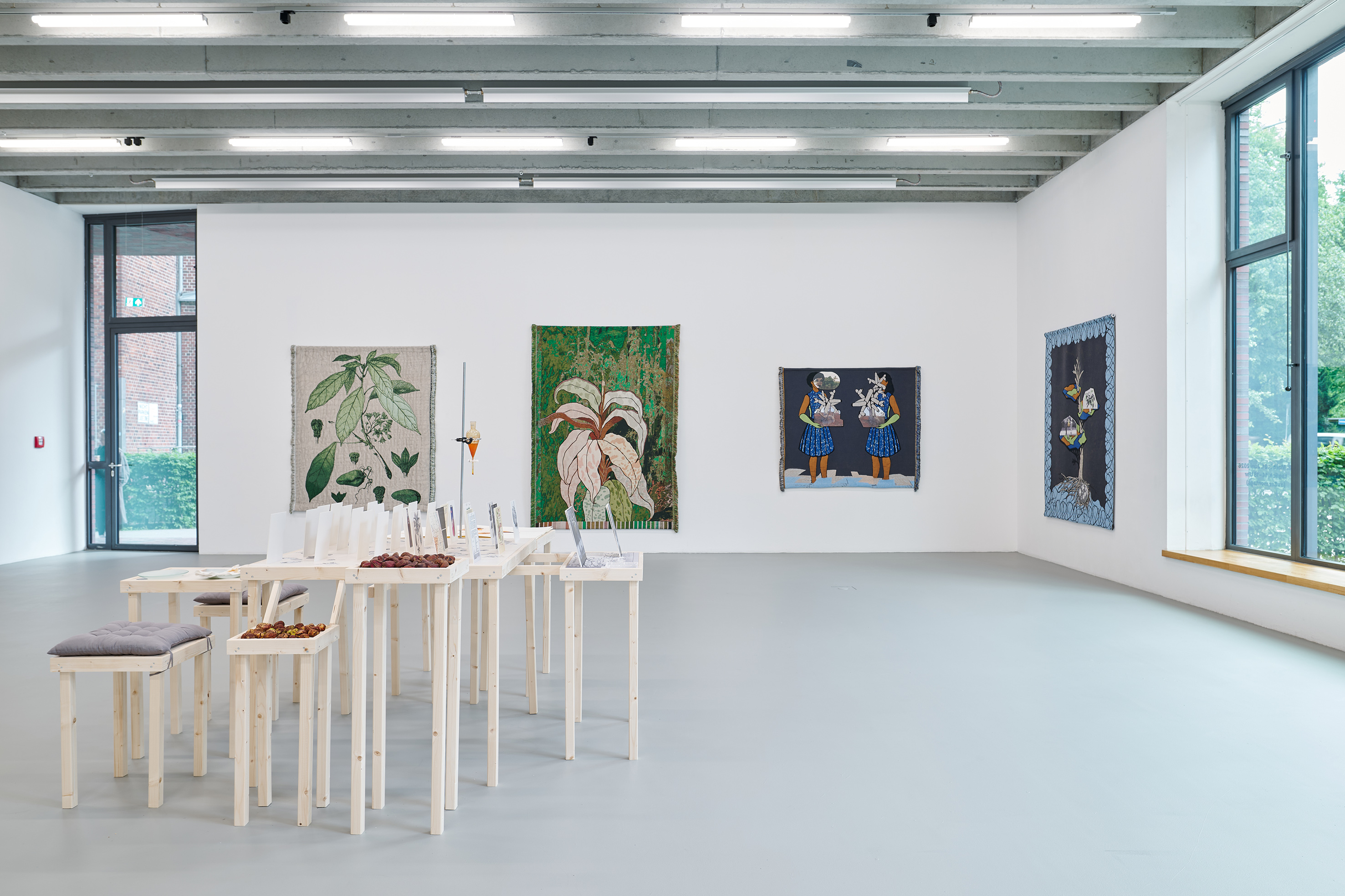A spacious, light-filled exhibition gallery features an installation of simple wooden tables and stools displaying printed materials, small objects, and plant-based elements. Several large-scale textile works with botanical motifs and figurative imagery hang on the white walls. Large windows and the open layout create a calm, research-oriented atmosphere.