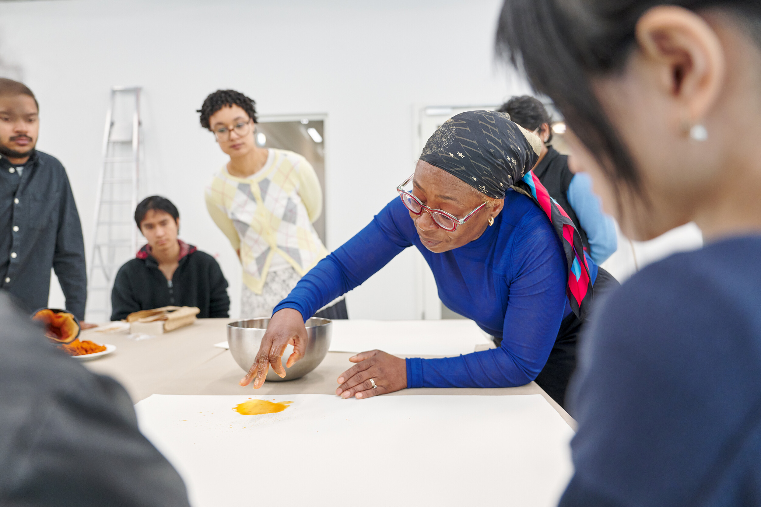 In a bright exhibition space, a group of people gathers around a large table. A person wearing a blue top and patterned headscarf demonstrates a hands-on process by applying a bright orange powder to a white surface with their fingers. The surrounding participants watch attentively. Bowls and materials on the table suggest a participatory workshop or a collaborative artistic practice.