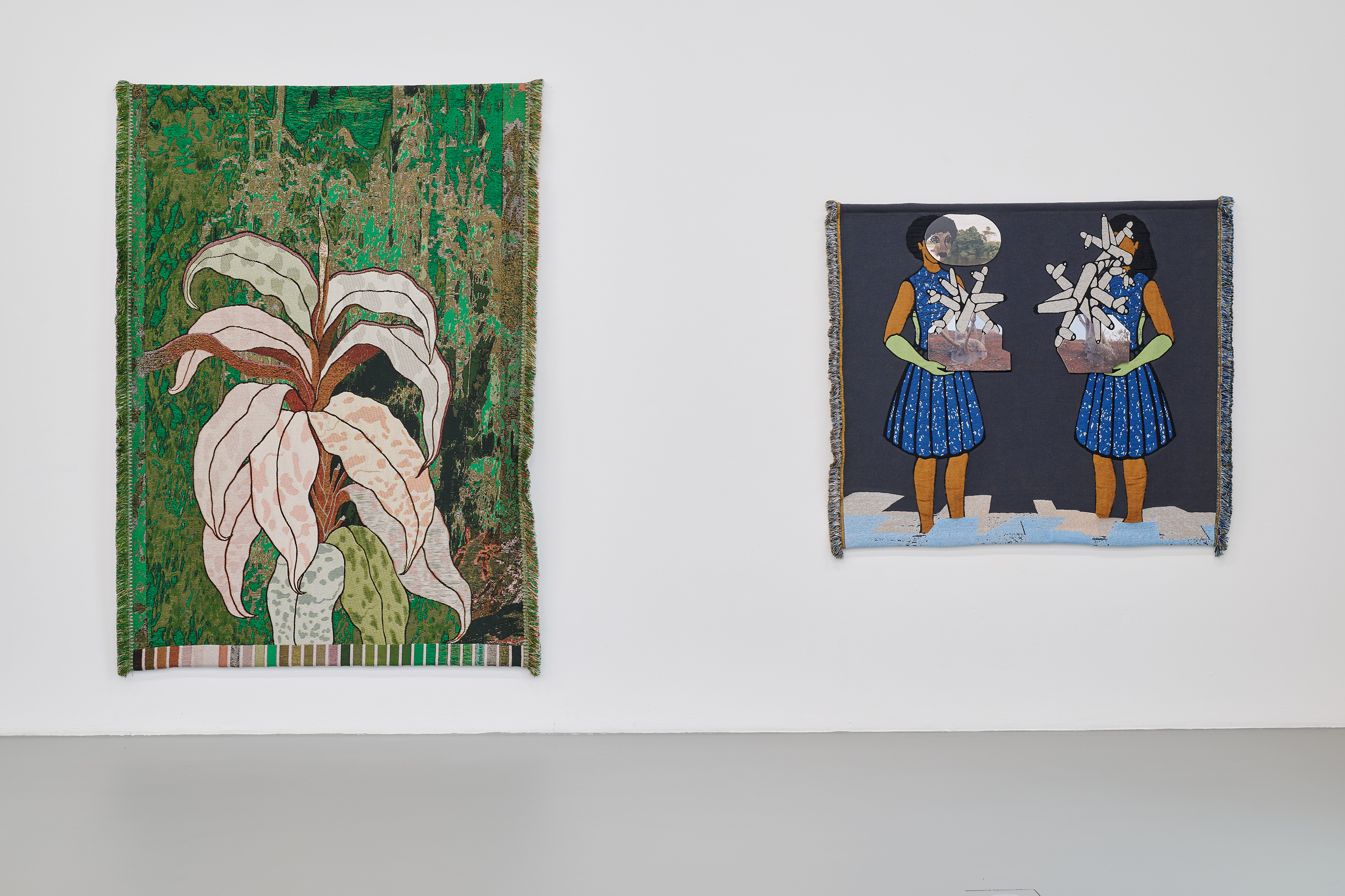 Two large-scale textile wall hangings are displayed on a white gallery wall. The work on the left depicts a stylized plant with long, pale leaves set against a richly textured green background. The piece on the right shows a figurative scene with two mirrored figures in blue dresses holding geometric objects in front of their bodies. Both works feature fringed edges and combine textile craftsmanship with pictorial and narrative imagery.