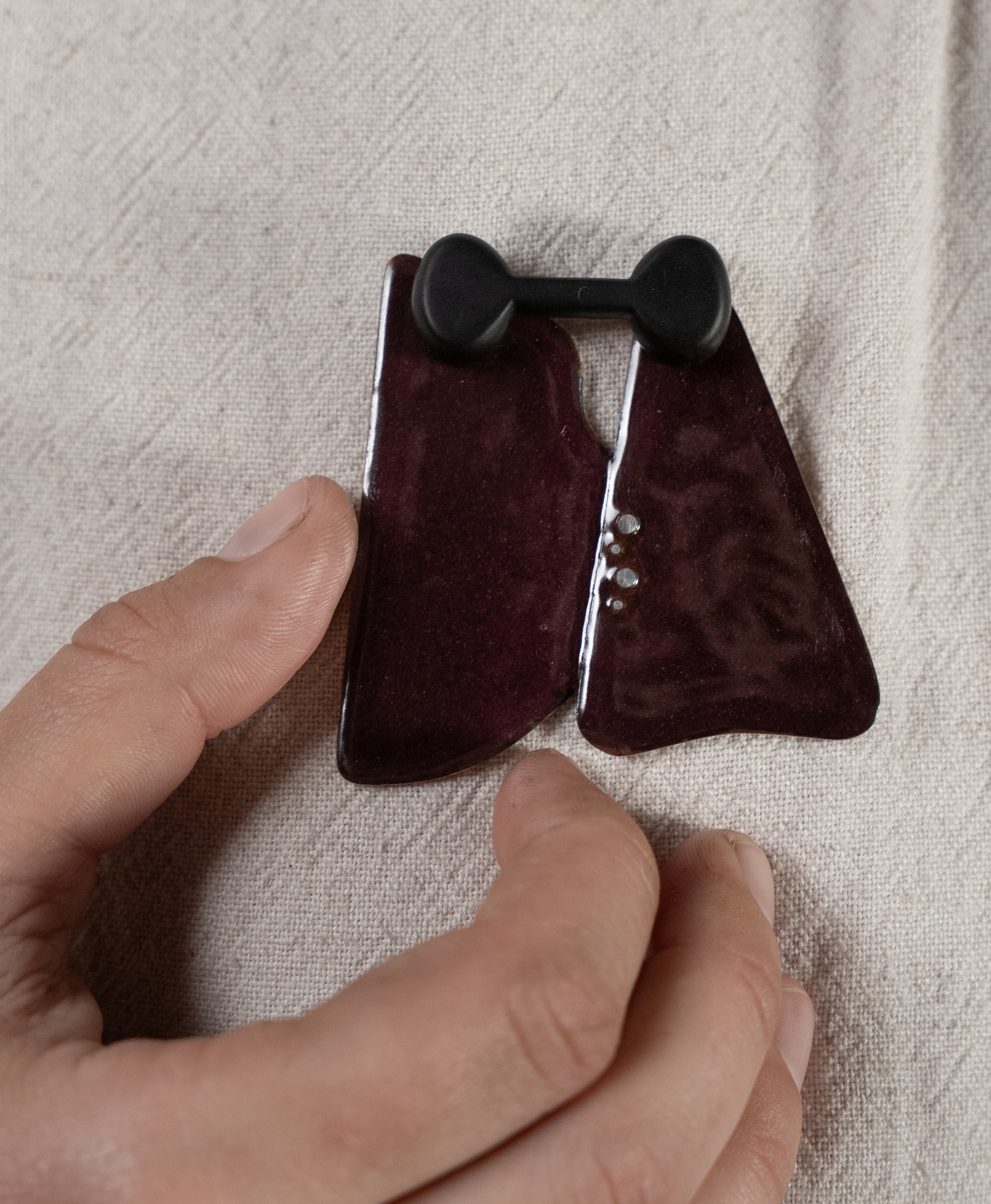 Nahaufnahme einer kleinen, dunkelroten Brosche in Form eines stilisierten Anzugs mit schwarzer Fliege, die auf hellem Stoff liegt und von einer Hand berührt wird.