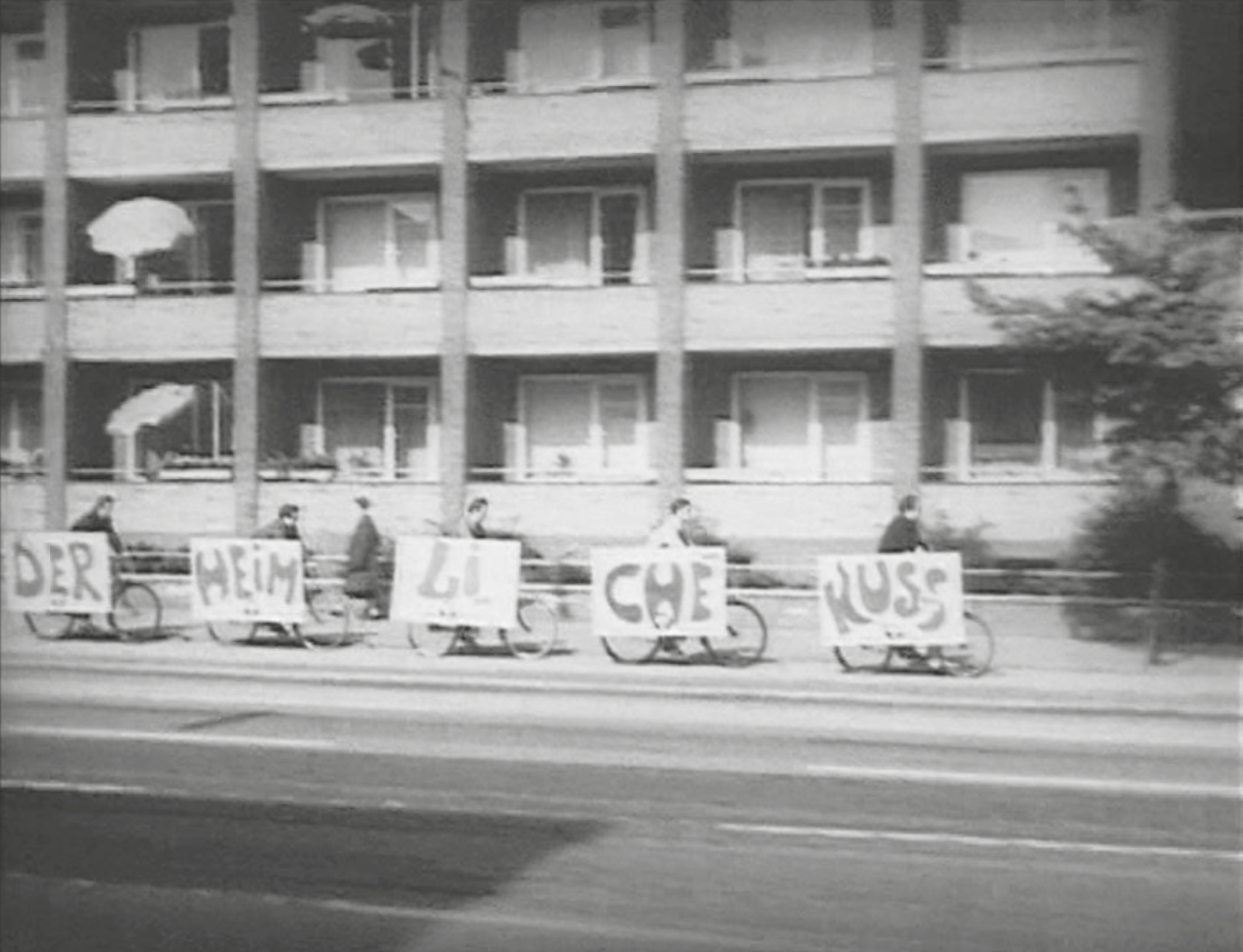 Black-and-white, slightly blurred photograph of a street action. Several people ride bicycles in a line, each carrying a large sign. The signs display individual words that together form a slogan. In the background, a multi-story residential building with balconies is visible.