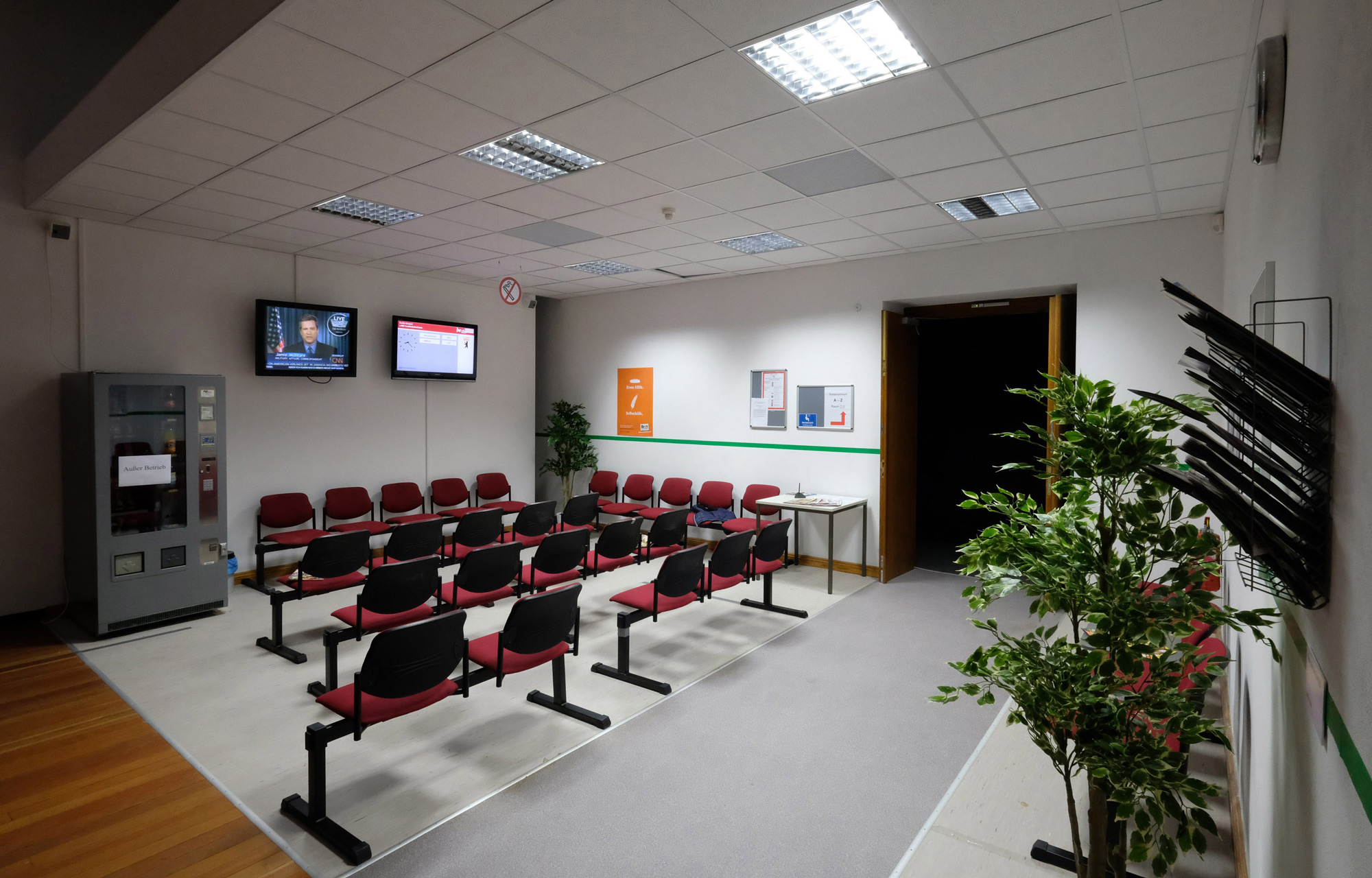 Waiting area with several rows of red and black chairs in a brightly lit room. Two televisions are mounted on the wall, alongside notices and a vending machine. A doorway leads into a darker adjacent room, and several plants are placed around the space.