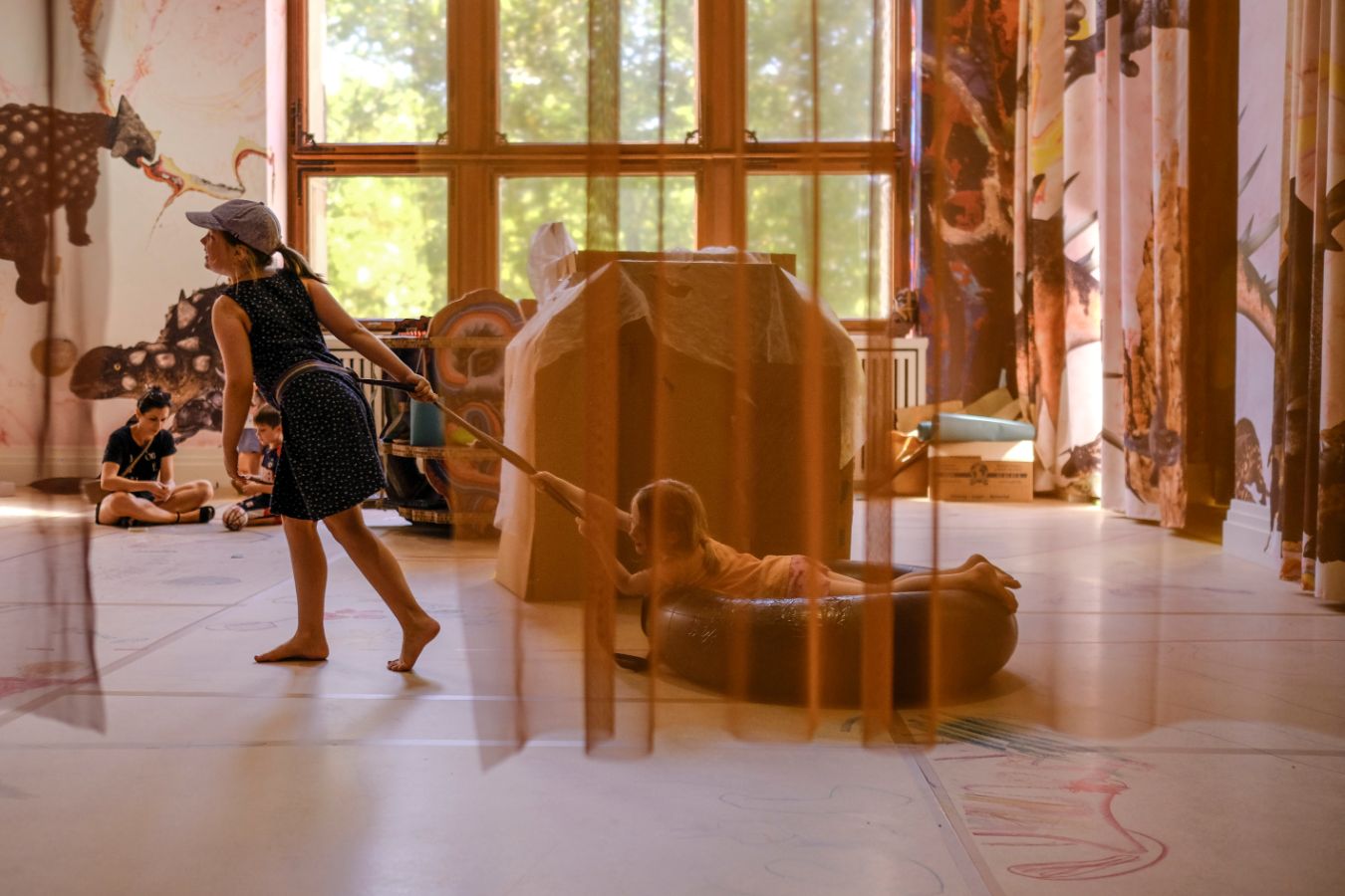 Several children are playing in a bright exhibition space with large windows. In the foreground, a girl wearing a cap pulls another child lying on their stomach in an inflatable swim ring across the floor. In the background, two children sit on the floor. Large illustrated curtains with animal motifs hang on the walls.