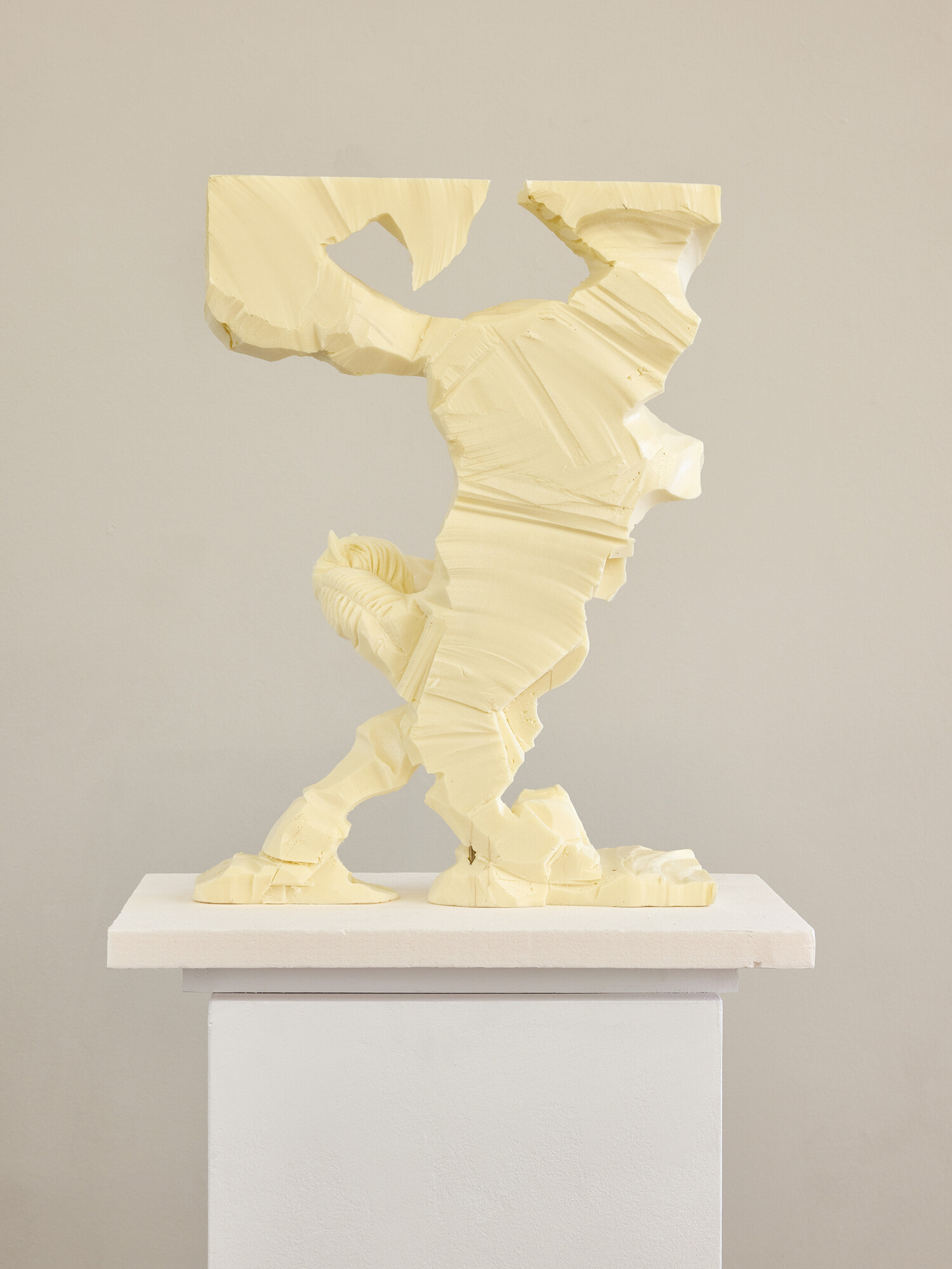 Eine helle, abstrakte Skulptur steht auf einem weißen Sockel vor einer weißen Wand. Die Form erinnert entfernt an eine menschliche Figur in Bewegung, wirkt jedoch fragmentiert und aus grob bearbeitetem Material herausgeschnitten. Sichtbare Schnitt- und Bearbeitungsspuren verleihen der Arbeit eine dynamische, skizzenhafte Anmutung zwischen Figuration und Abstraktion.