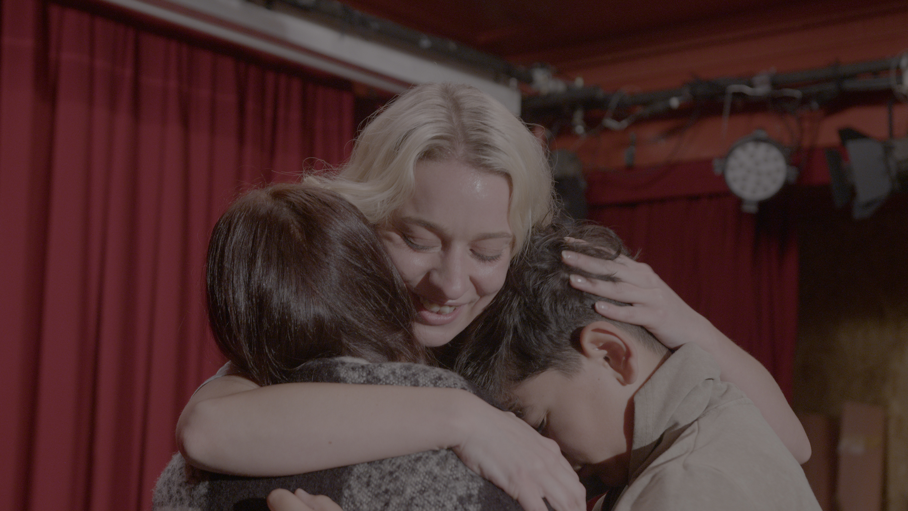 Three people share a close embrace in a theatre or performance space. At the center, a smiling blonde person wraps their arms around the other two, who rest their heads against their shoulders. Red stage curtains and lighting equipment are visible in the background. The scene conveys intimacy, comfort, and a sense of connection.