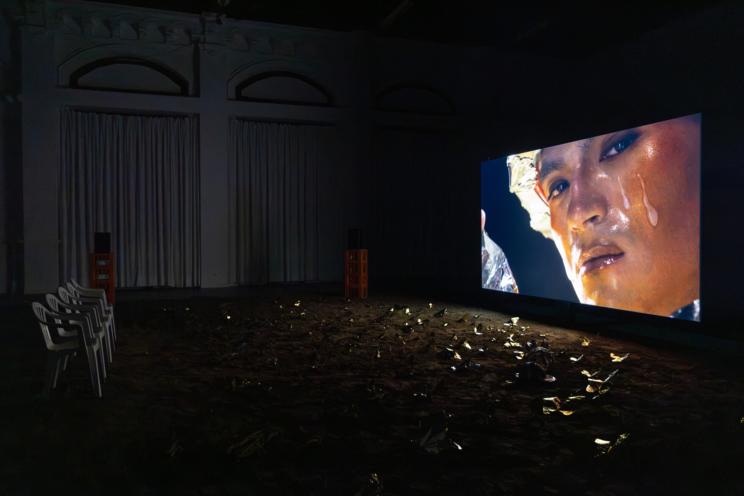 Eine raumfüllende Videoinstallation in einem abgedunkelten Ausstellungsraum. Auf einer großen Leinwand ist die Nahaufnahme eines Gesichts zu sehen, über das Flüssigkeit läuft. Vor der Projektion stehen mehrere weiße Stühle; der Boden ist mit dunklem Material oder Objekten bedeckt, die das Licht der Projektion reflektieren. Die Installation erzeugt eine immersive, filmische Atmosphäre.