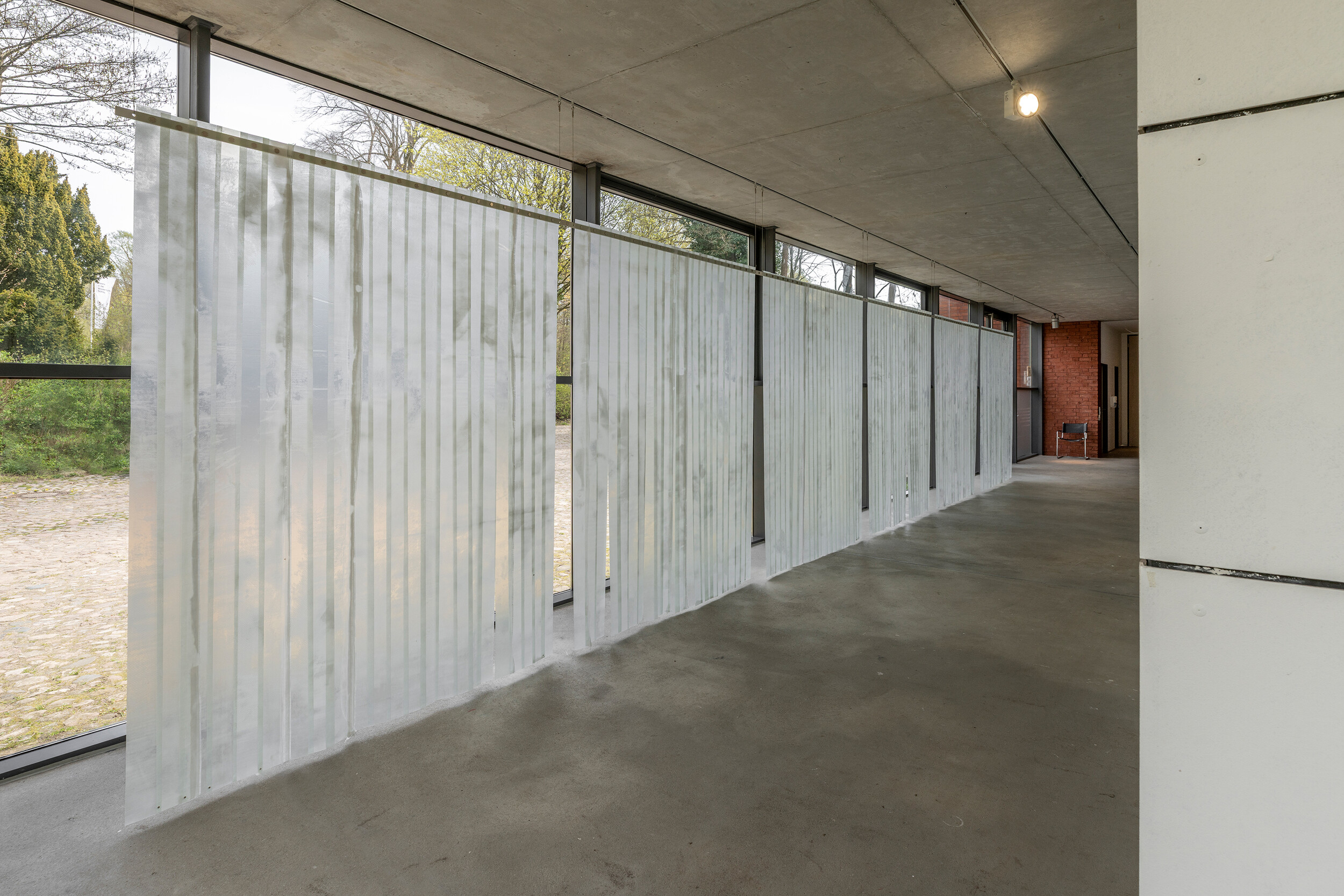 In einem langgestreckten, modernistischen Korridor mit bodentiefen Fenstern erstreckt sich eine raumgreifende Installation aus halbtransparenten, geriffelten Kunststoffbahnen. Die vertikal hängenden Elemente bilden eine durchlässige Wand entlang der Fensterfront und verändern die Lichtverhältnisse im Raum. Durch das milchige Material bleiben Außenraum und Architektur nur schemenhaft sichtbar.