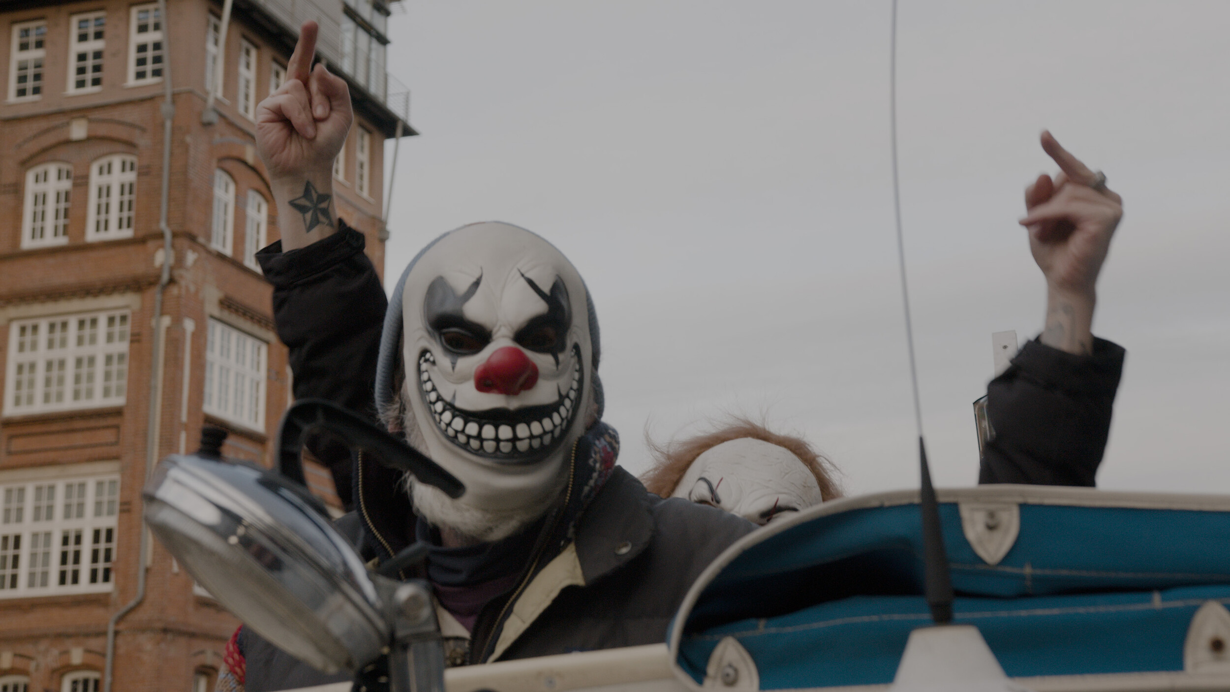 Eine Person mit einer grinsenden Clownsmaske sitzt in einem offenen Fahrzeug und hebt einen ausgestreckten Mittelfinger in die Luft. Eine zweite maskierte Person ist im Hintergrund teilweise sichtbar und macht dieselbe Geste. Hinter ihnen erhebt sich ein Backsteingebäude vor einem grauen Himmel. Die Szene wirkt provokativ und rebellisch.