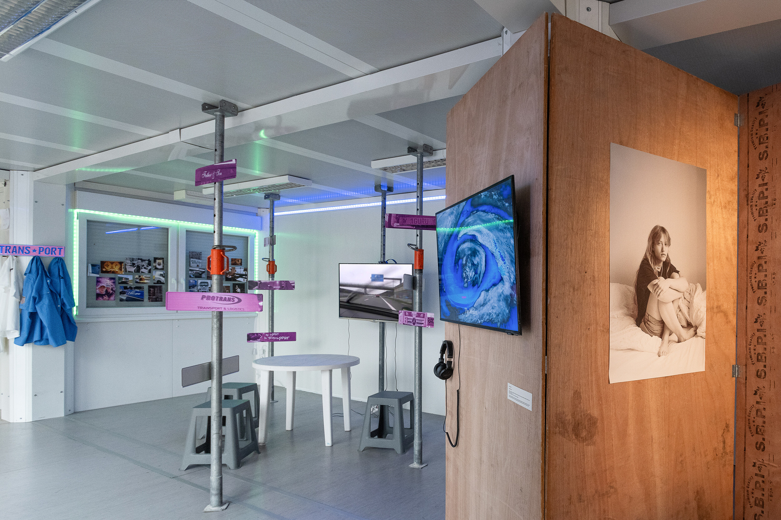 Eine multimediale Installation in einem hellen, modularen Ausstellungsraum kombiniert Video, Fotografie, Text, Objekte und grafische Elemente. Monitore zeigen bewegte Bilder, während farbige Beschriftungen und industrielle Stützen den Raum strukturieren. Ein runder Tisch mit Hockern lädt zum Verweilen ein. Im Vordergrund ist eine Holzwand mit einem großformatigen Schwarz-Weiß-Porträt einer sitzenden Person zu sehen. Neonartige Lichtleisten verleihen der Installation eine technisch anmutende Atmosphäre.