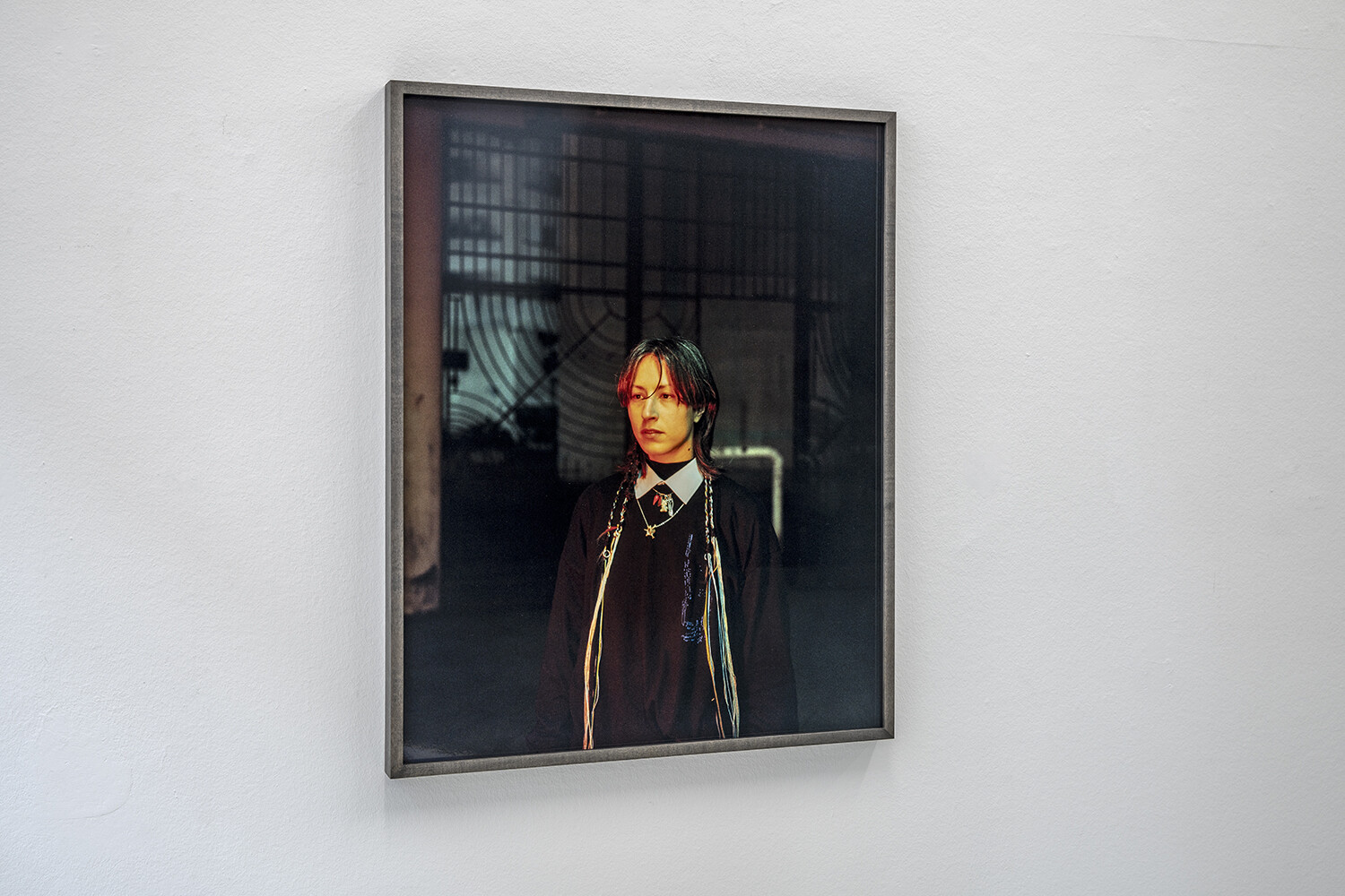 A framed colour photograph is mounted on a white gallery wall. The image depicts a person dressed in dark clothing against a dimly lit background. Warm light illuminates the face, while geometric structures and reflections in the background create an atmospheric and contemplative mood.