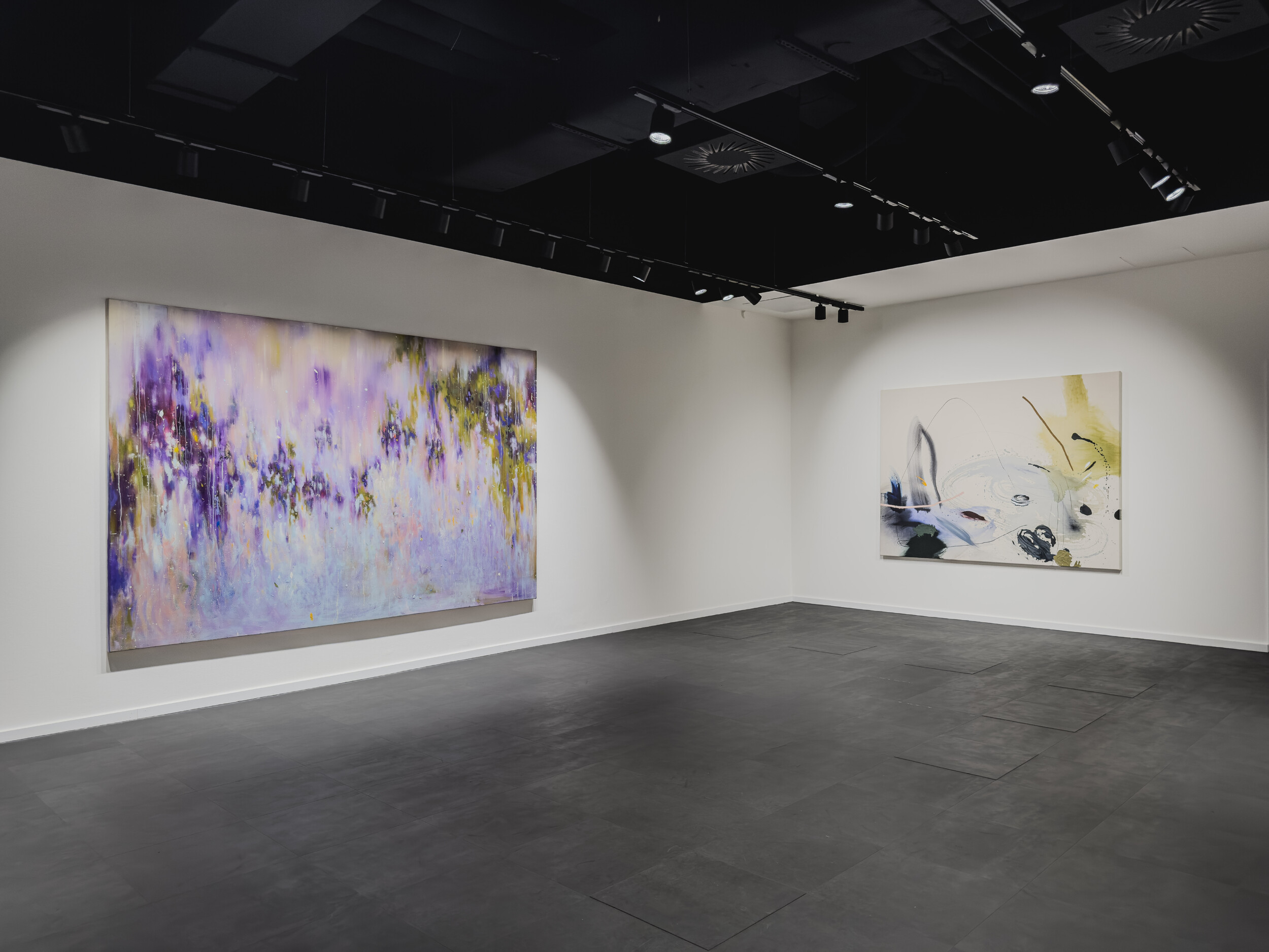 Ein heller Ausstellungsraum mit dunklem Boden und weißen Wänden zeigt zwei großformatige abstrakte Gemälde. Links hängt eine überwiegend in Violett-, Rosa- und Gelbtönen gehaltene Malerei mit fließenden, atmosphärischen Farbverläufen. Rechts befindet sich ein helles Bild mit gestischen Linien, schwarzen und farbigen Akzenten sowie offenen, skizzenhaften Formen. Die Werke werden durch gerichtete Deckenstrahler beleuchtet.