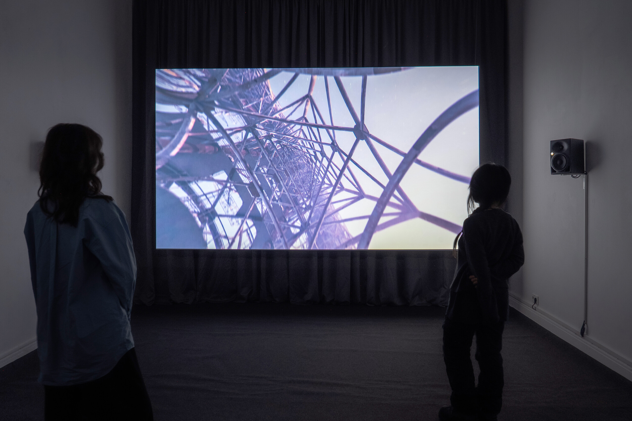 Zwei Personen betrachten in einem abgedunkelten Ausstellungsraum eine großformatige Videoprojektion, die den Blick durch eine komplexe metallene Gitterkonstruktion zeigt.