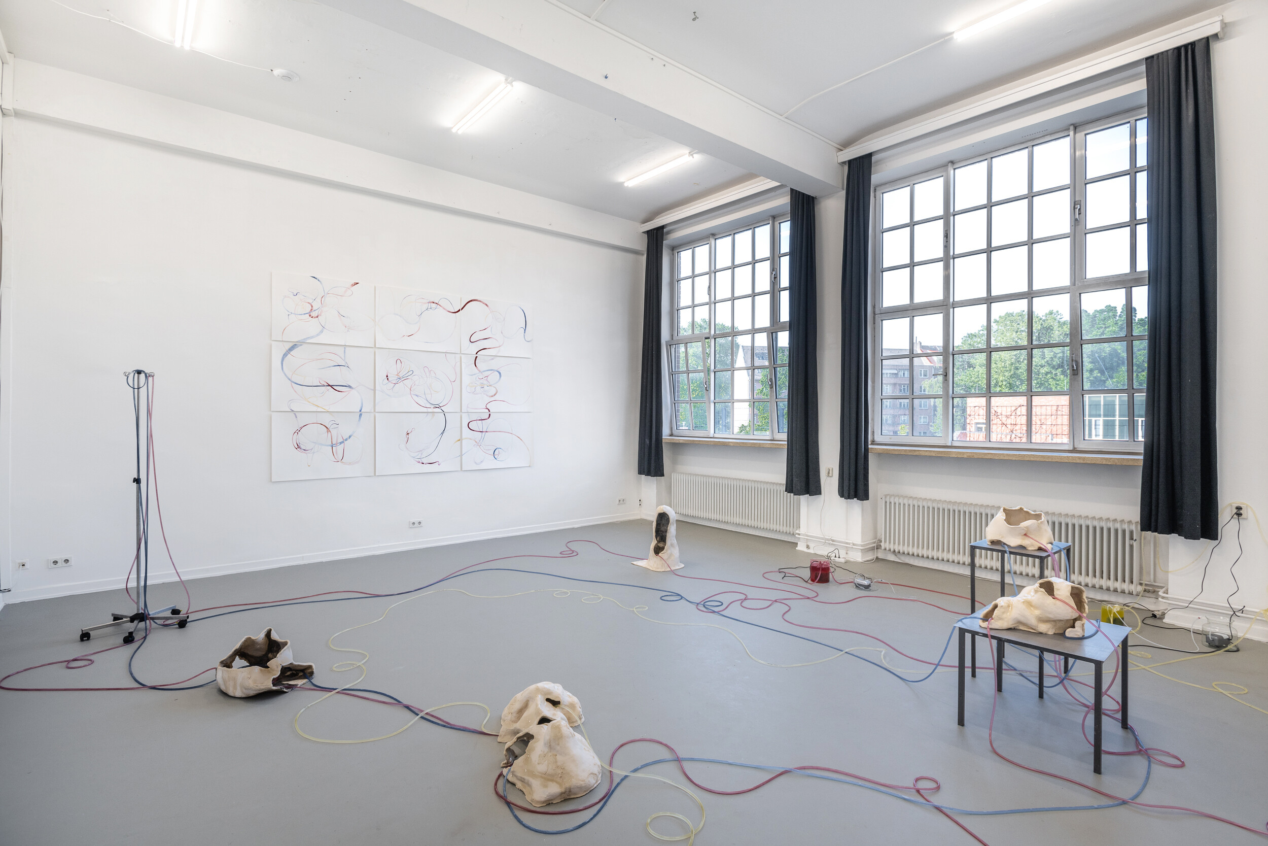 Ausstellungsansicht in einem hellen Raum mit großen Fenstern: Organisch geformte, helle Skulpturen liegen auf dem Boden und auf Metallgestellen. Farbige Kabel verbinden sie miteinander und führen zu einer wandfüllenden Zeichnung aus geschwungenen Linien.