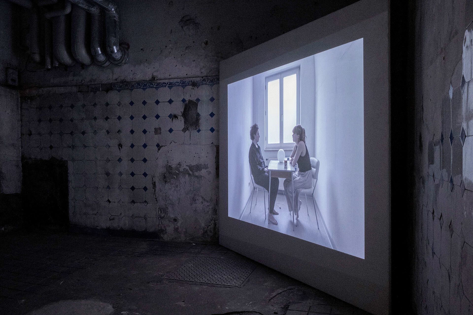 Videoprojektion in einem dunklen, verfallenen Raum mit beschädigten Fliesen und freiliegenden Rohren. Das projizierte Bild zeigt zwei Personen, die sich in einem hellen Raum an einem kleinen Tisch gegenübersitzen.