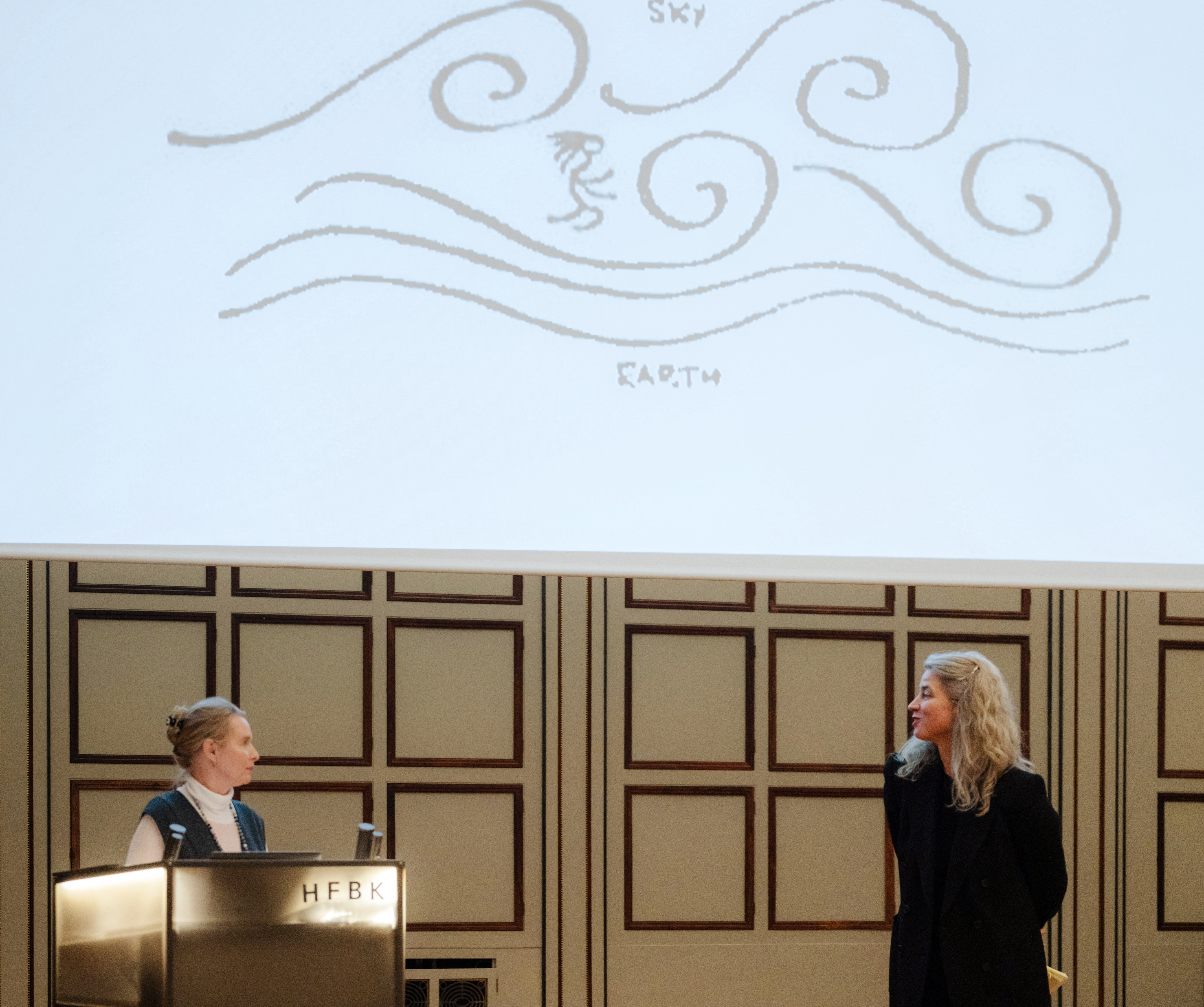 Zwei Frauen unterhalten sich im Stehen auf einem Podium, über ihren Köpfen ist eine Projektion von gezeichneten Luftströmungen zu sehen.