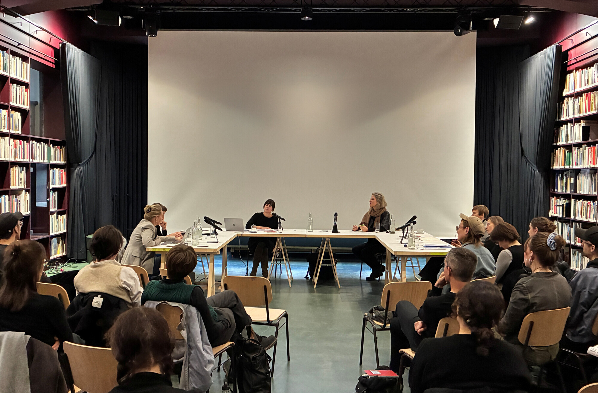 Mehrere Frauen sitzen an einem in U-Form aufgestellten Tisch und diskutieren vor einem sitzenden Publikum. Links und rechts sind seitlich in mehreren Reihen viele Bücher untergebracht.