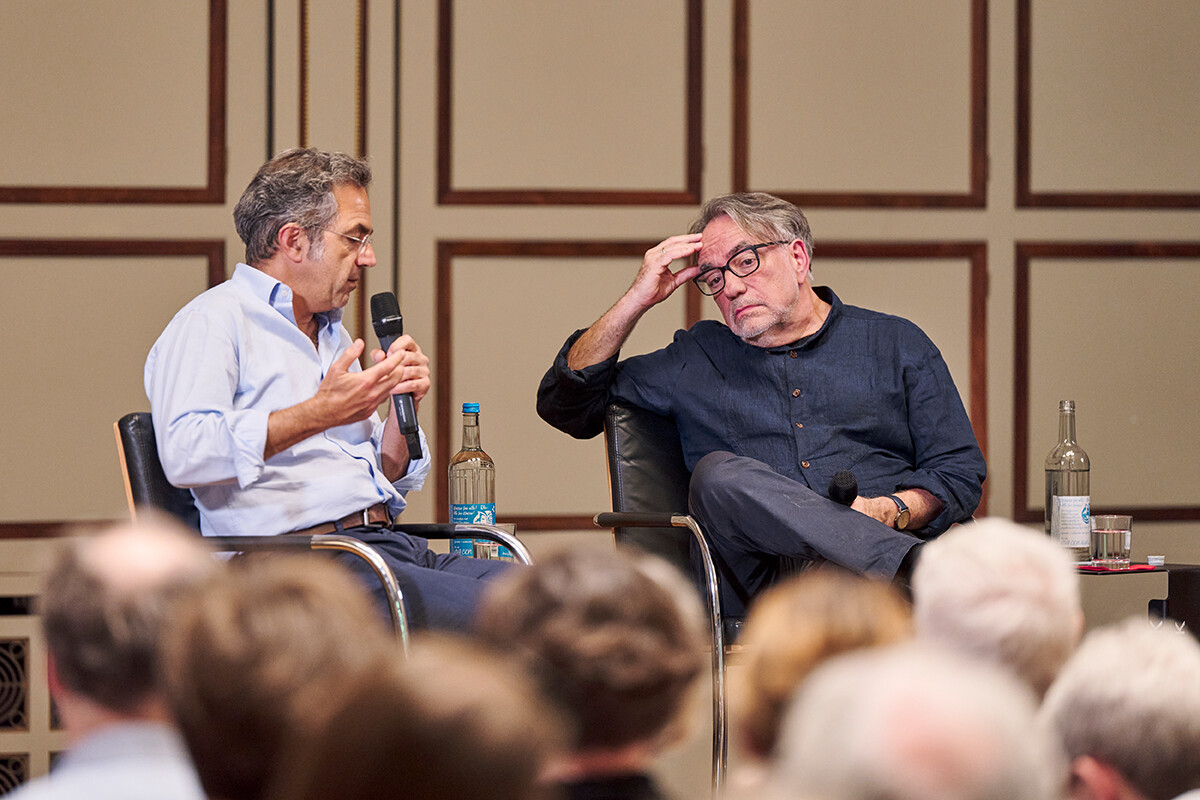 Ein Mann mit einem Mikrophon spricht, ein zweiter hört angestrengt zu.