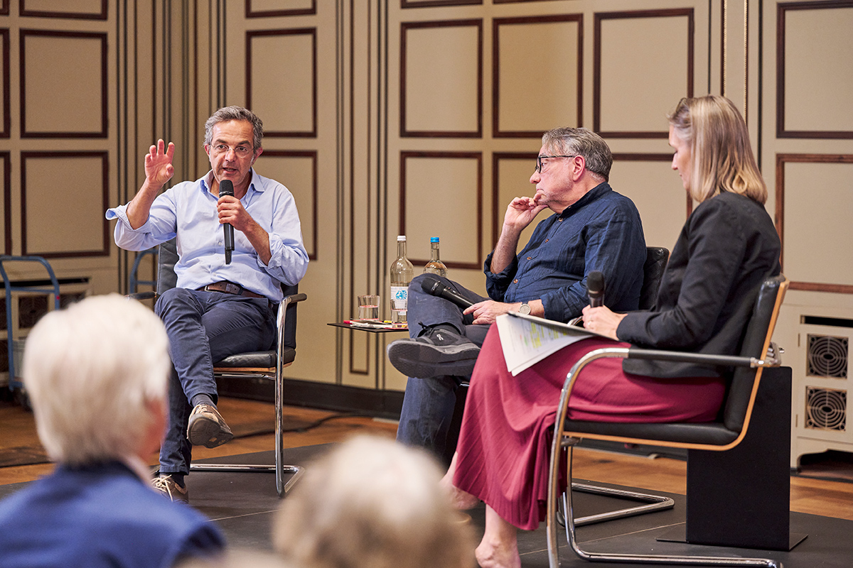 Zwei Männer und eine Fraue sitzen auf einem Podium im Gespräch
