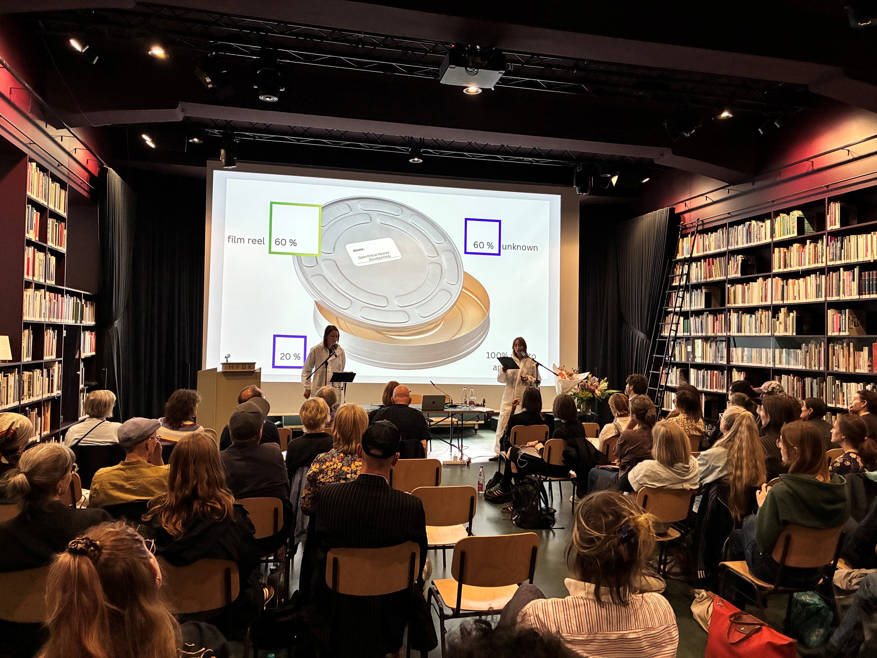 Blick in die Extended Library der HFBK Hamburg: Zwei Vortragende stehen vor einem großen Publikum. Hinter ihnen ist eine Präsentation mit einer Filmrolle und grafischen Elementen auf eine Leinwand projiziert.
