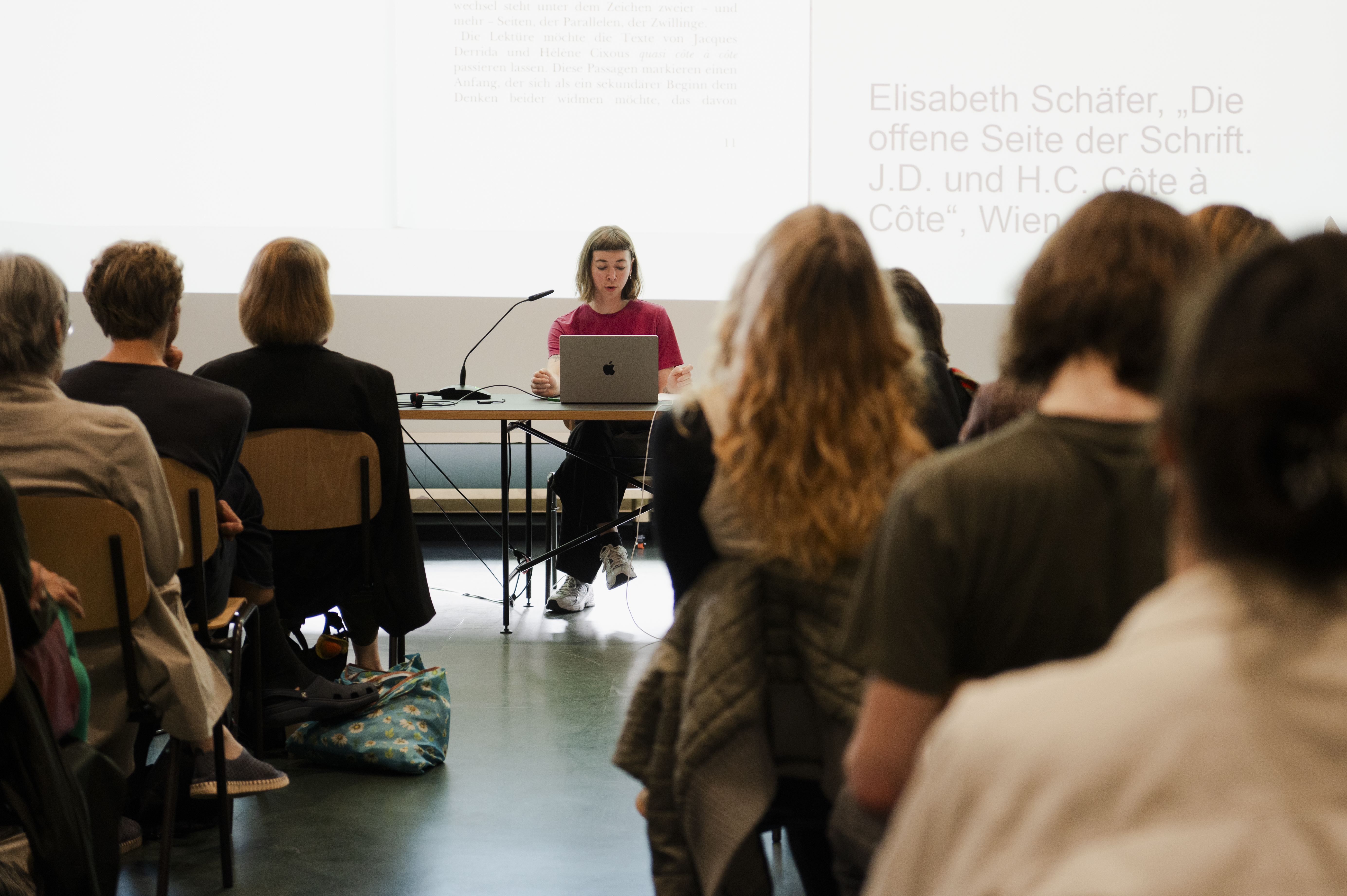 Blick in die Extended Library der HFBK Hamburg: Eine Referentin sitzt an einem Tisch mit Laptop und spricht vor Publikum. Hinter ihr ist eine Präsentation mit projiziertem Text zu sehen.