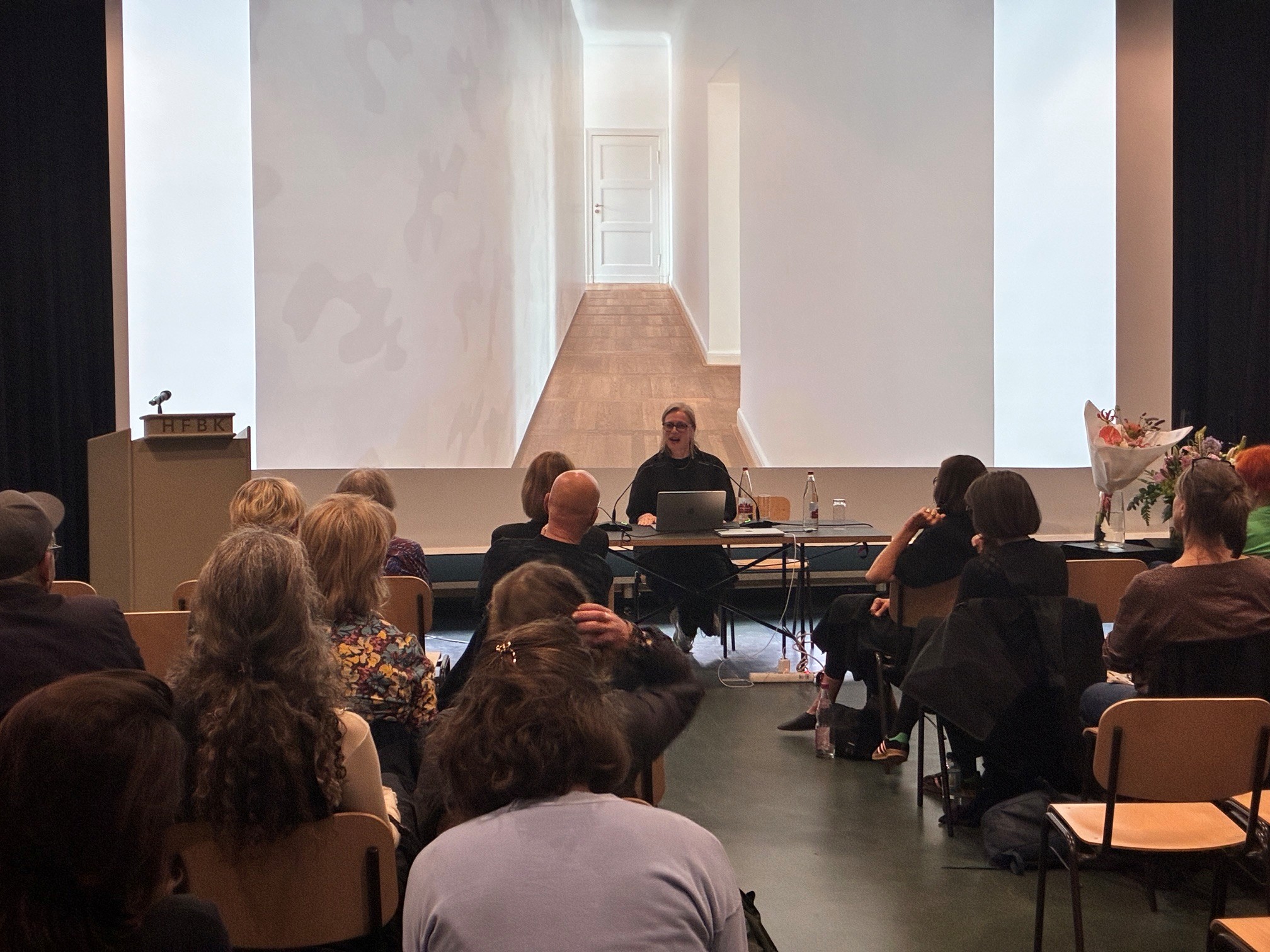 Blick in die Extended Library der HFBK Hamburg: Eine Referentin spricht vor Publikum, während hinter ihr eine großformatige Projektion eines Innenraums mit Flur und Tür zu sehen ist.