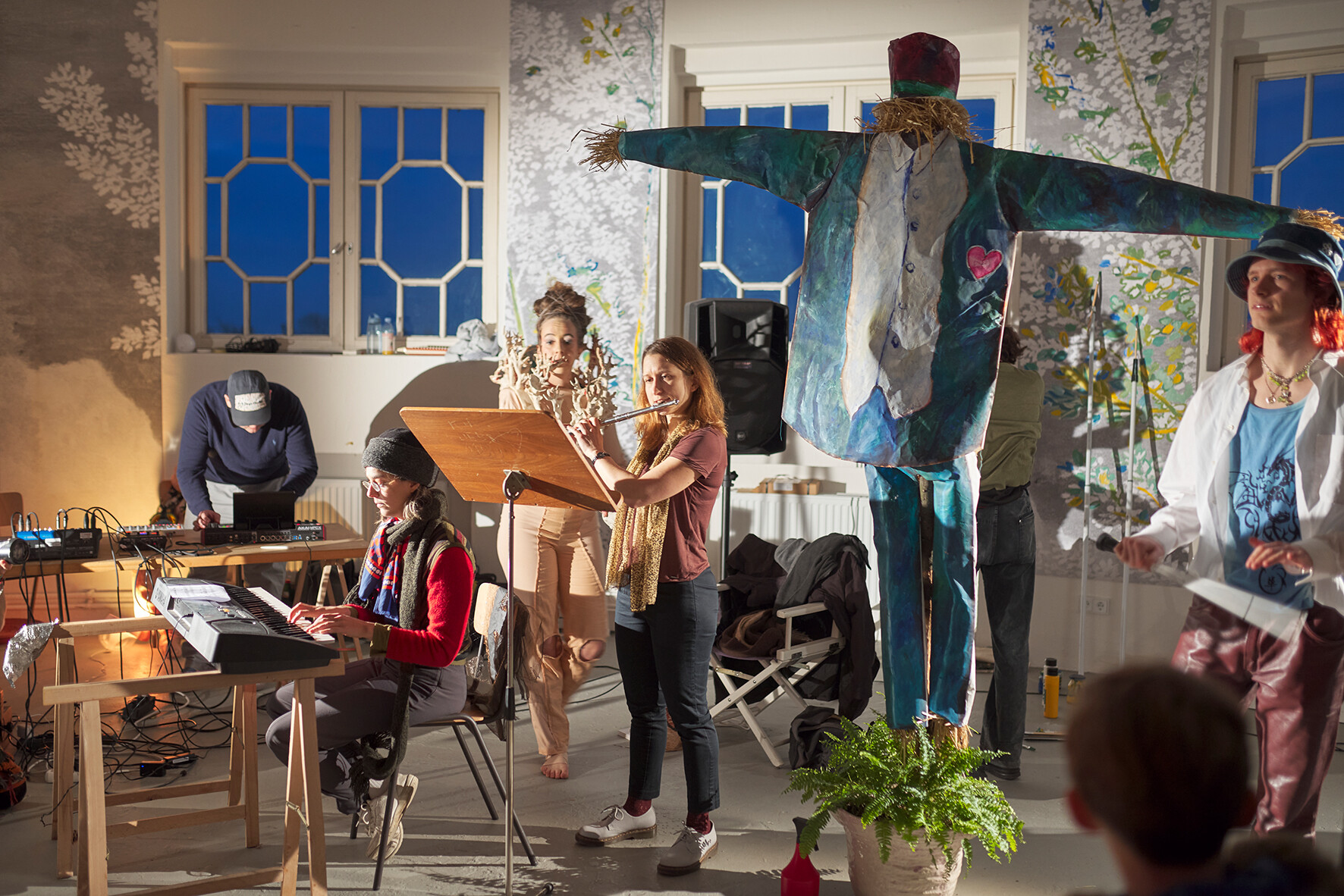 Artistic performance in a bright room with several participants. One person plays a keyboard while another plays the flute, as others in the background handle technical equipment or props. A large painted scarecrow figure with outstretched arms stands in the room. The warm lighting creates a theatrical atmosphere.