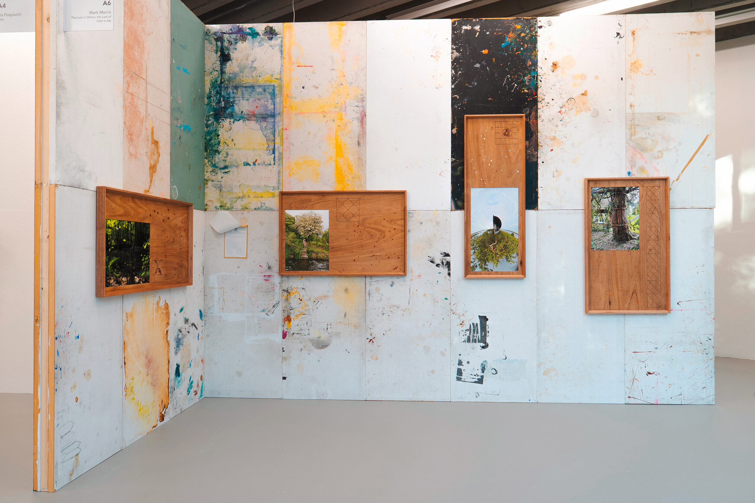 Ausstellungsansicht mit vier gerahmten Naturfotografien auf einer aus gebrauchten, farbverschmierten Platten zusammengesetzten Stellwand. Die Holzrahmen weisen eingravierte Muster und kleine Bohrungen auf.