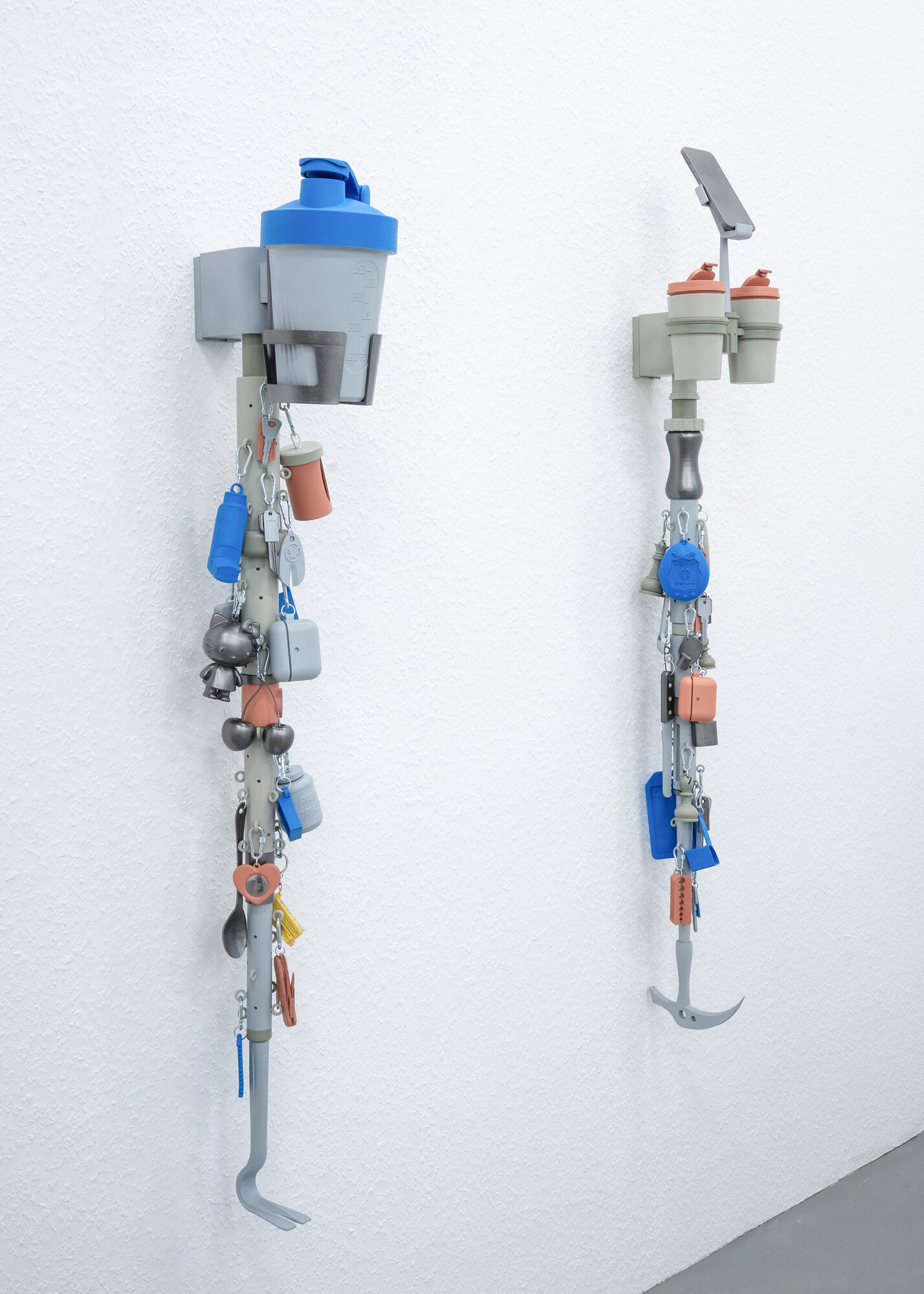 Zwei vertikale, an der Wand befestigte Assemblagen aus Trinkbehältern, Halterungen, Schlüsseln, Anhängern, Karabinerhaken und Werkzeugen in Grau, Blau und Orange.
